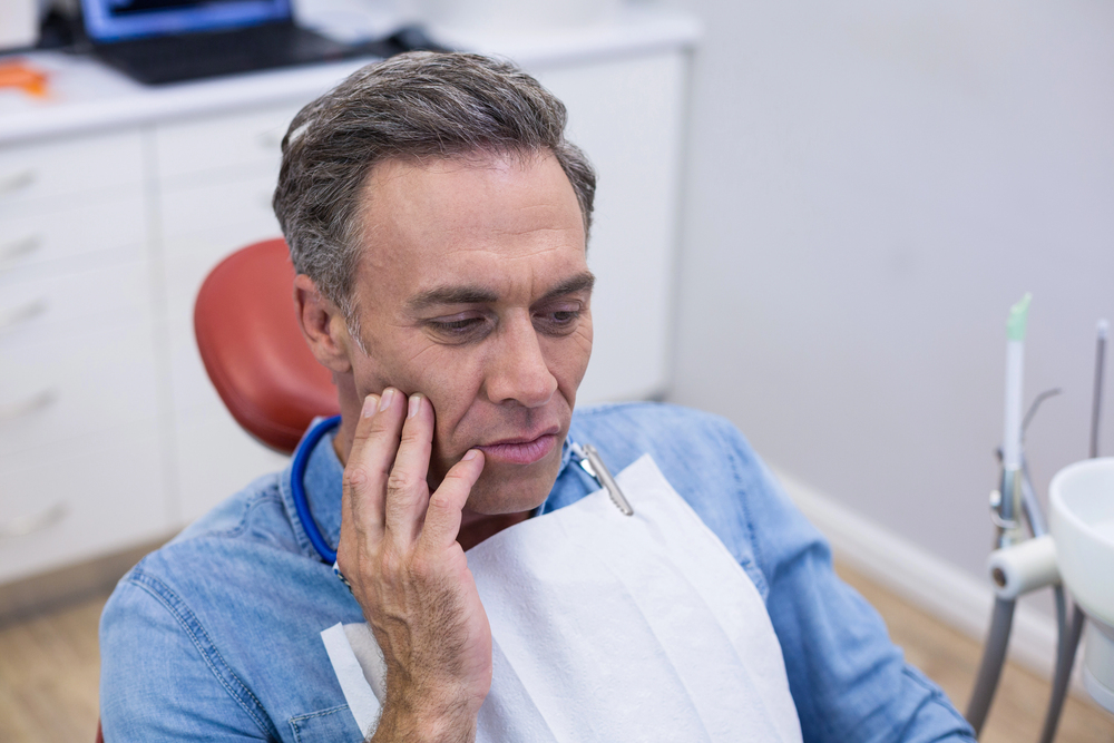 older man touching his face in pain, sitting in dental chair, fear of the dentist