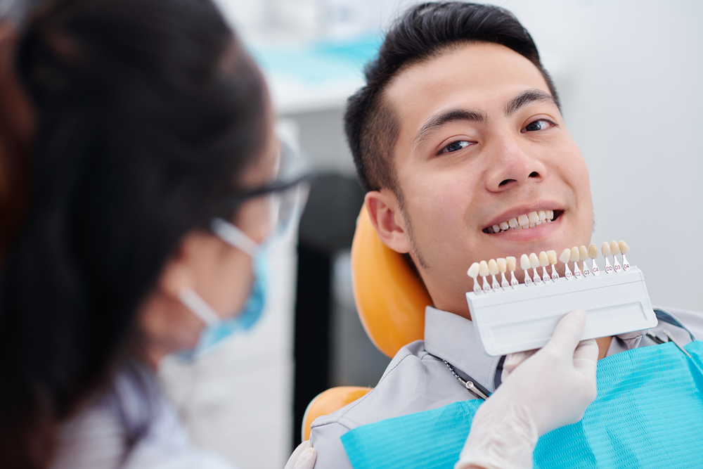 Dentist using special palette when choosing teeth color of smiling male patient before whitening procedure