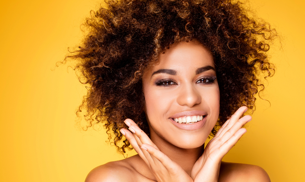 black woman holding her hands in a v-shape under her chin on yellow background